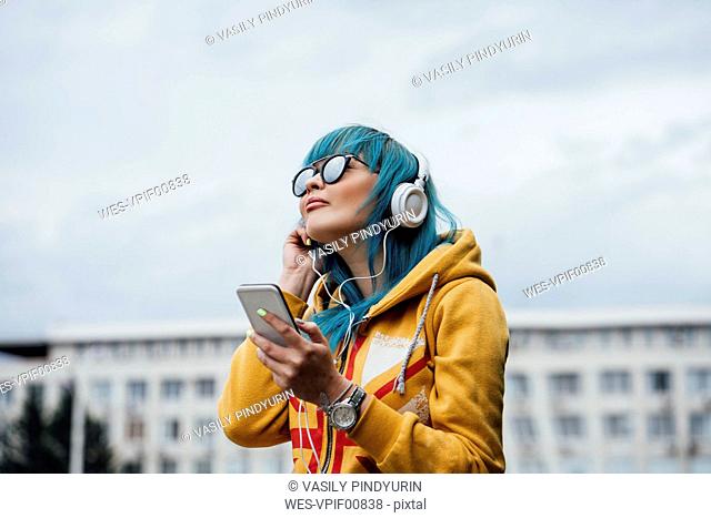 Portrait of young woman with dyed blue hair listening music with smartphone and headphones