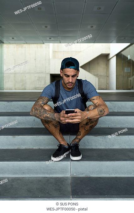 Tattooed young man sitting on stairs using smartphone