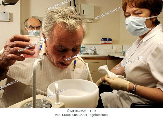 An elderly man rinsing his mouth out with anti-microbial solution, watched by the dentist and his assistant
