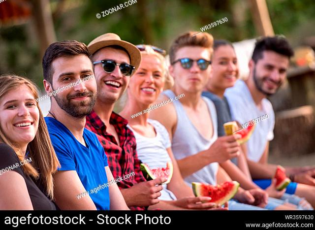 group of young friends enjoying watermelon while sitting on the wooden bridge over the river in beautiful nature