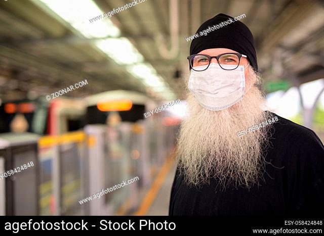 Portrait of mature bearded hipster man as tourist with mask for protection from corona virus outbreak at the sky train station