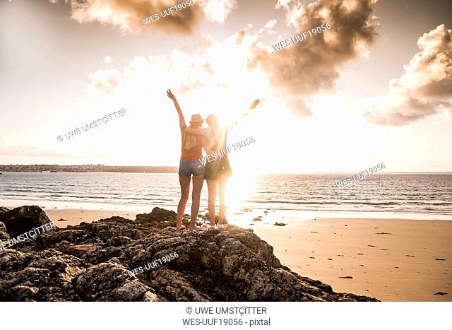 Two girlfriends standing on rocky beach, waving at sunset, rear view