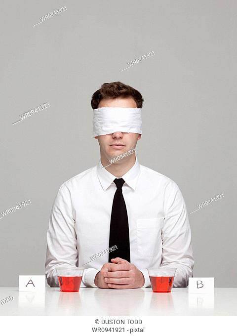 Studio portrait of young man with blindfold sitting in front of two glasses with red liquid