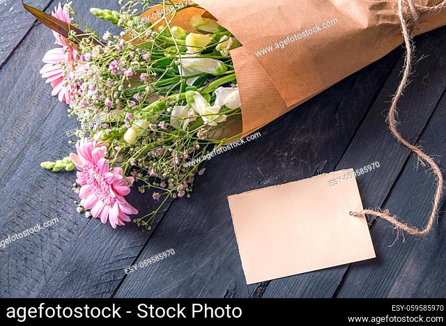 Greeting card idea with a cheerful bouquet of flowers wrapped in vintage brown paper and an empty etiquette tied to it, on a rustic wooden table