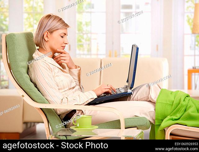 Woman sitting in armchair with feet up working with computer at home in living room