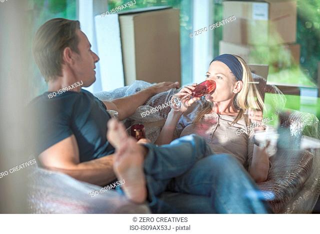 Moving house: couple relaxing on bubbled wrapped sofa, in room with cardboard boxes, drinking from champagne flutes