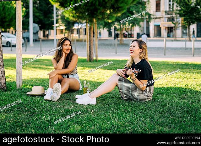Young woman looking at carefree friend playing ukulele while sitting on grassy land