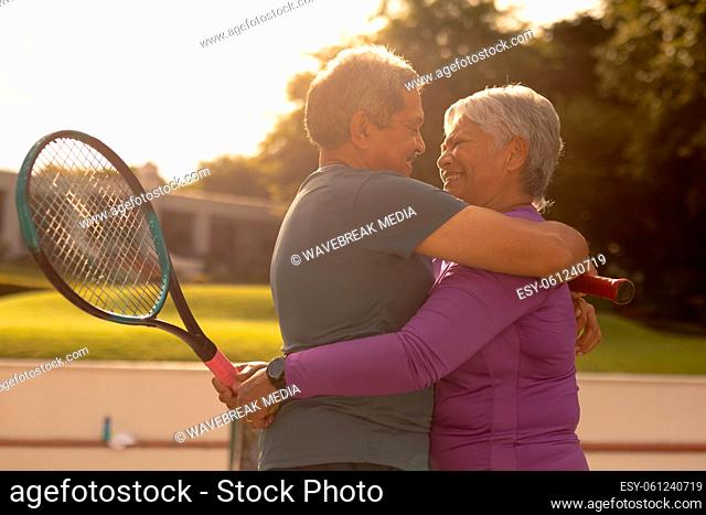 Cheerful romantic biracial senior couple embracing while standing at tennis court during sunset