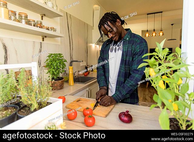 Smiling young man cutting yellow chili pepper in kitchen