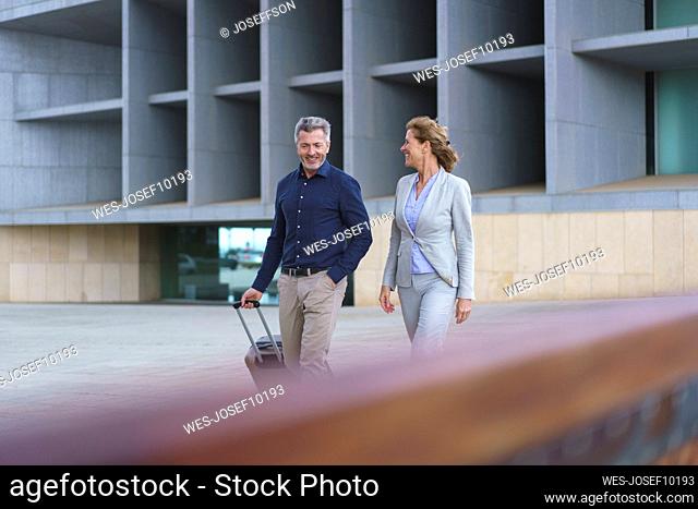 Smiling businessman with suitcase walking by businesswoman in front of office building