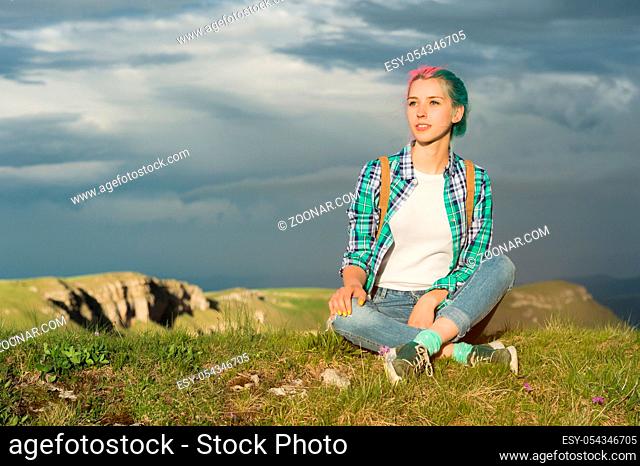Portrait of young smiling woman traveler with multi-colored hair. Sitting high in the mountains in the evening with a decline in the background of a plateau