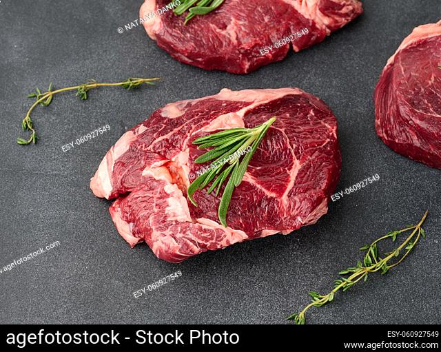 raw piece of beef ribeye with rosemary, thyme on a black table, top view