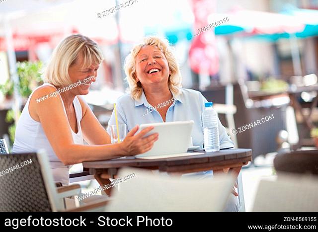 Two female friends using touch pad in street cafe and laughing. Senior women spending enjoyable summer day