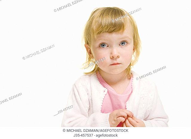 A three year old girl with blonde hair wearing a white sweater and a pink top looks into camera with a concerned expression while holding her hands together