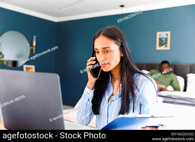 Biracial woman using laptop and talking on smartphone working in bedroom, with partner in background