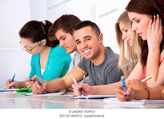 Handsome Student Sitting With Classmates Writing At Desk