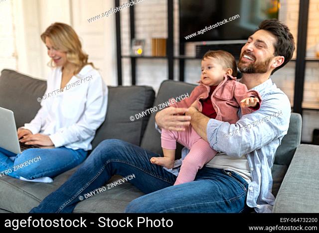 Father carrying sleeping daughter while looking at woman using laptop on sofa