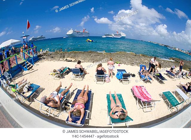 Tourists sunning on beach chairs on the waterfront in Georgetown on Grand Cayman in the Cayman Islands with cruise ships in the caribbean sea off shore taken...