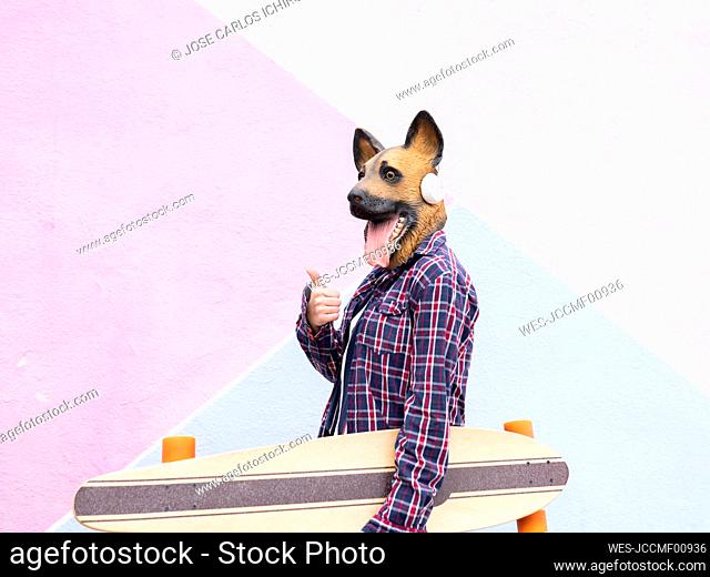 Young woman wearing dog mask showing thumbs up while holding skateboard against wall