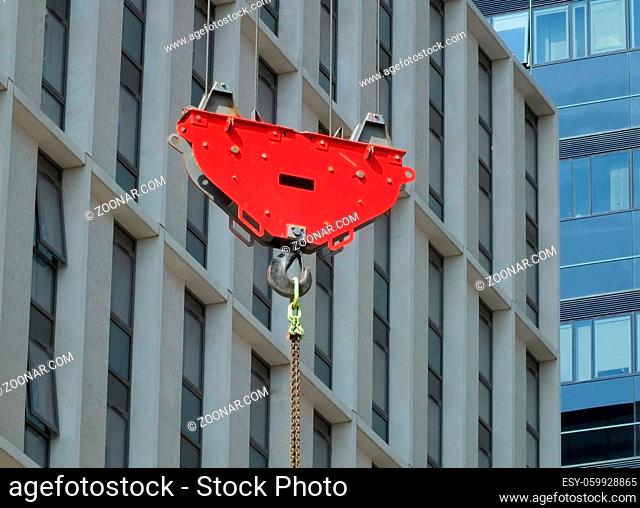 a bright red crane hook and pulley lifting a suspended metal chain on a construction site with urban modern buildings in the background