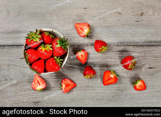 Tabletop view - small ceramic bowl of strawberries, more cut fruits lying around on gray wood desk