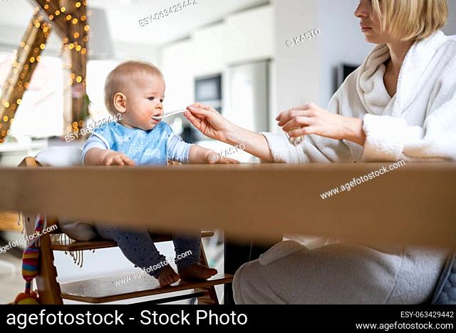 Mother wearing cosy bathrope spoon feeding her baby boy child in baby high chair with fruit puree at dinning table at home