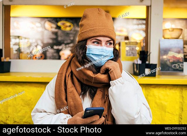 Young woman with hand on chin day dreaming while holding smart phone against cafe during COVID-19
