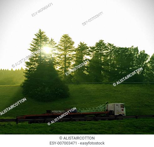 Sun and forest trees shine down on long semi truck delivery with motion blur on highway road