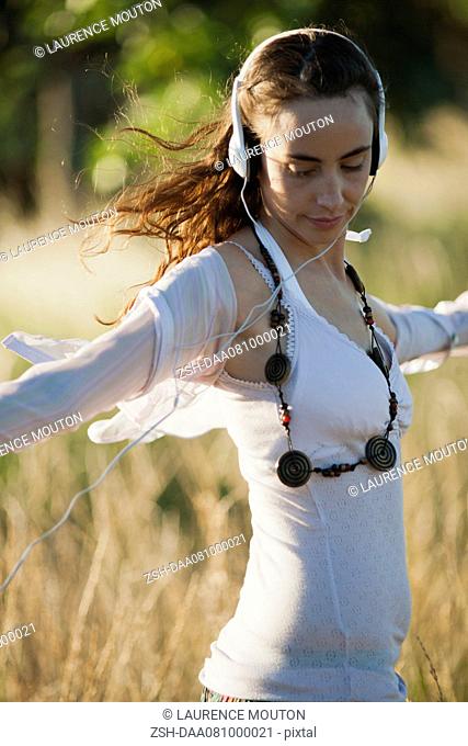 Young woman listening to headphones in field with arms outstretched