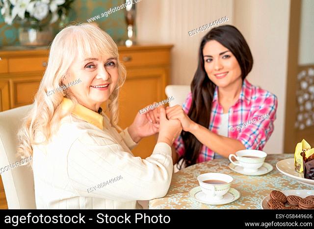 Selective Focus On Happy Elderly Woman Holding Hands With Her Younger Friend At The Dining Table. Blurred Portrait Of Daughter Holding Hands With Mother