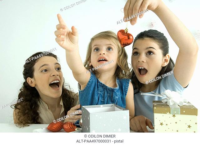 Girls opening presents, one holding ornament, other reaching up, mother watching
