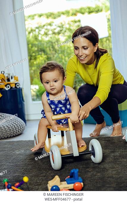 Happy mother with baby daughter driving toy car in living room