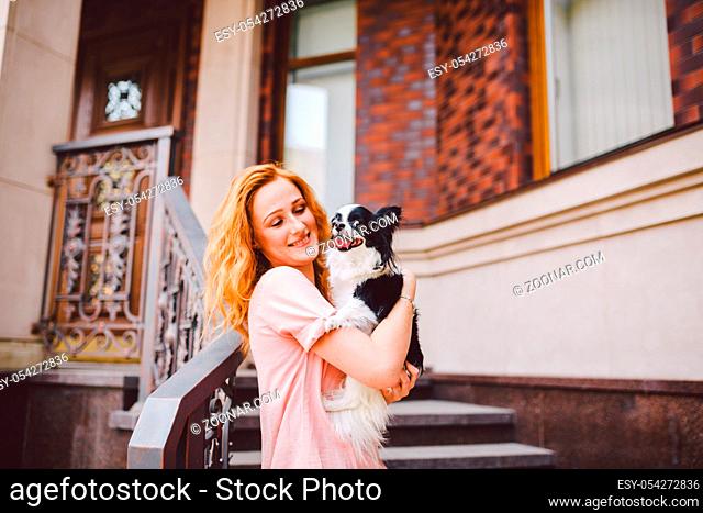 A beautiful young woman with red long hair is holding a small, cute funny big-eyed dog of two flowers, a black-and-white pet of the breed of hichuahua against a...