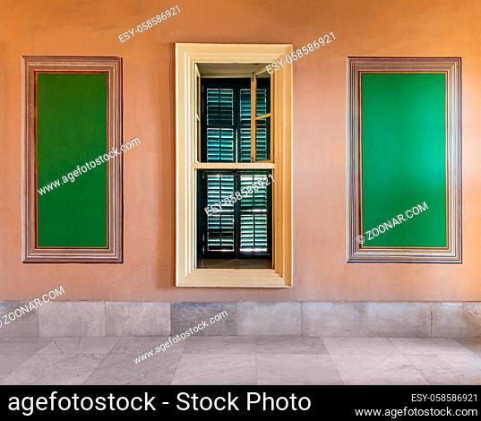 Narrow wooden window with closed green shutters mediating two beautiful elegant rectangular green frames on orange wall with white marble floor