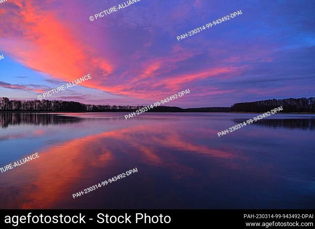 13 March 2023, Brandenburg, Altfriedland: Colorful clouds glow in the light of the sunset over the Klostersee lake in the district of Märkisch-Oderland