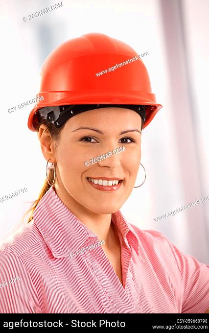 Closeup portrait of female architect wearing red hardhat, looking at camera, smiling