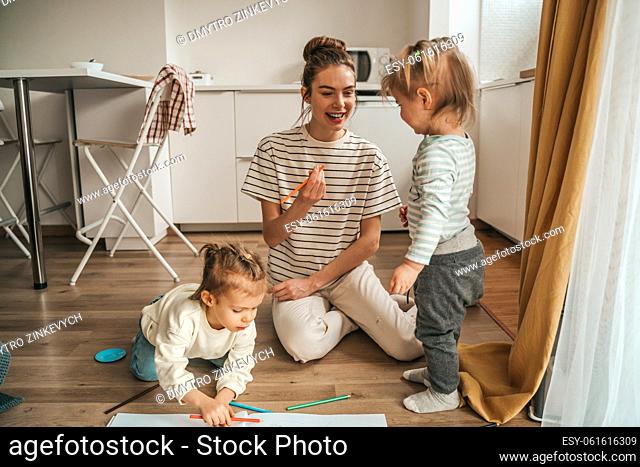 Joyous young mother with a colored pencil in the hand sitting on the kitchen floor among her children