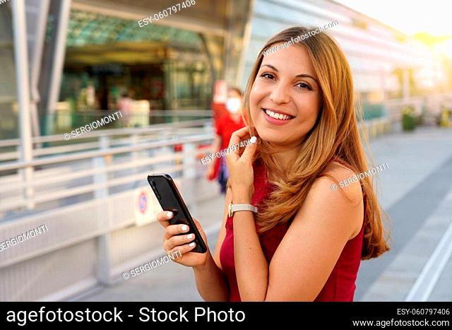 Young female model with long hair looks at the camera holding a mobile phone outdoors
