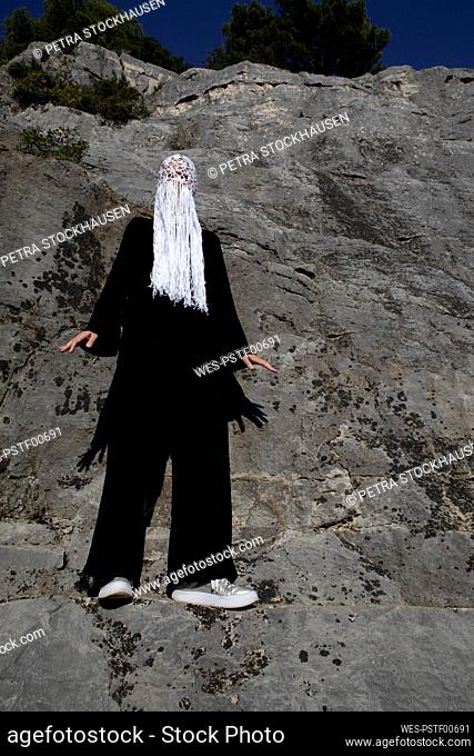 Woman dressed in black wearing crocheted white headdress with fringes standing in front of rock face