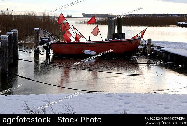 01 December 2023, Mecklenburg-Western Pomerania, Ahrenshoop: A snow-covered fishing boat lies in the Bodden harbor, which is already partially covered with a...