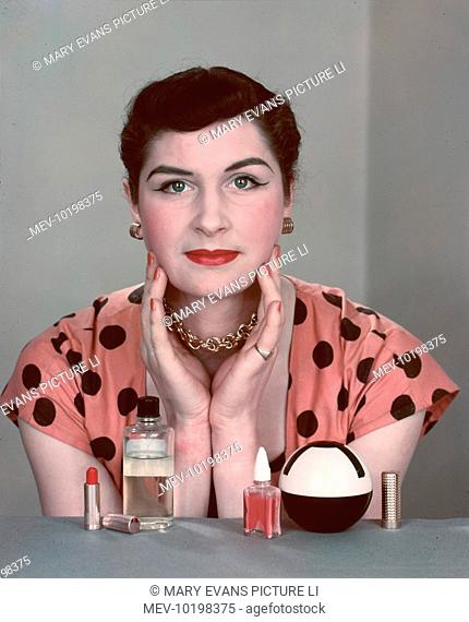 A brunette woman in a pink and black spotted dress sits at her dressing table going through her beauty routine.  Products include nail varnish