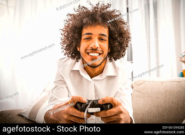 Arabic gay playing computer game. Young man holds Play Station joystick sitting at sofa in backlit. Smiling handsome looking at camera