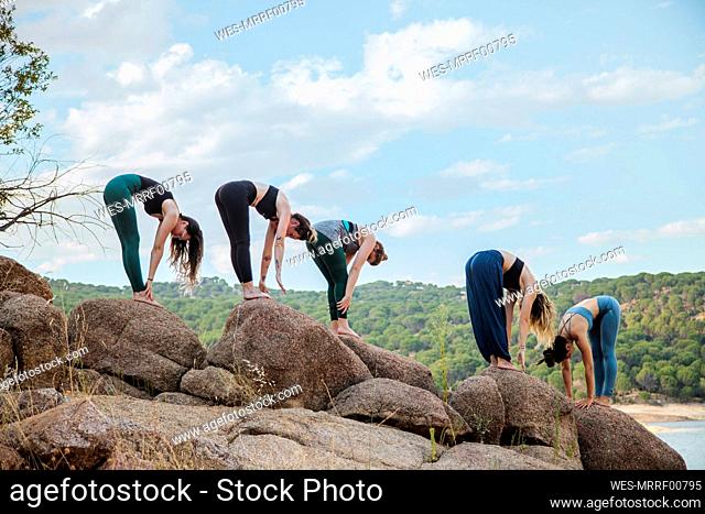 Young women with instructor practicing Uttanasana yoga on rock