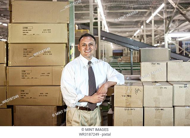 Portrait of a male Hispanic American executive in a shirt and tie surrounded by products stored in cardboard boxes in a large distribution warehouse