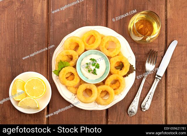 Squid rings with a mayonnaise dip, lemons, and a glass of wine, shot from the top on a dark rustic wooden background with copy space