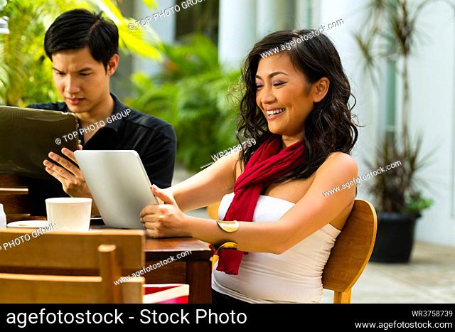 Asian woman and man are sitting in a bar or cafe outdoor and are surfing the internet with a tablet computer