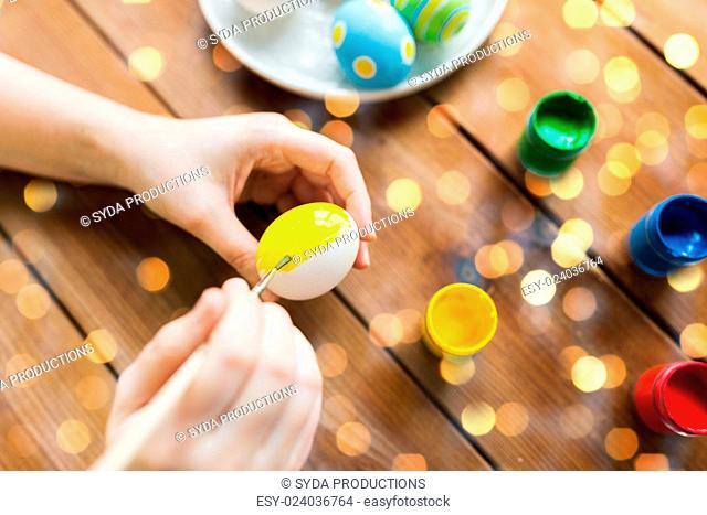 easter, holidays, tradition and people concept - close up of woman hands coloring easter eggs with colors and brush over holidays lights