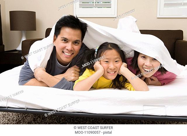 Mother father and daughter lying down on bed covered with bed sheet