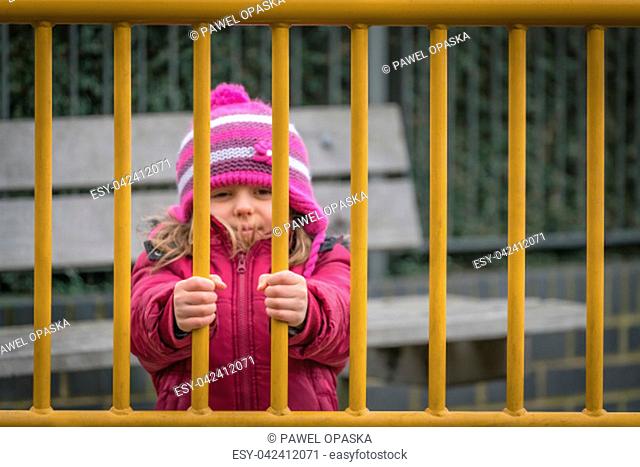Little cute Caucasian girl dressed in pink hat and jacket standing behind metal gates leading to the playground