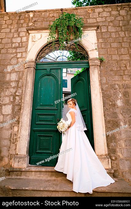 A gentle bride stands on the step and at the old green open door and holds a bouquet . High quality photo